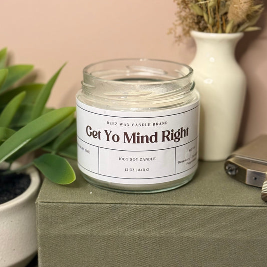 Candle jar labeled 'Get Yo Mind Right' on a surface with plants and a vase in the background.
