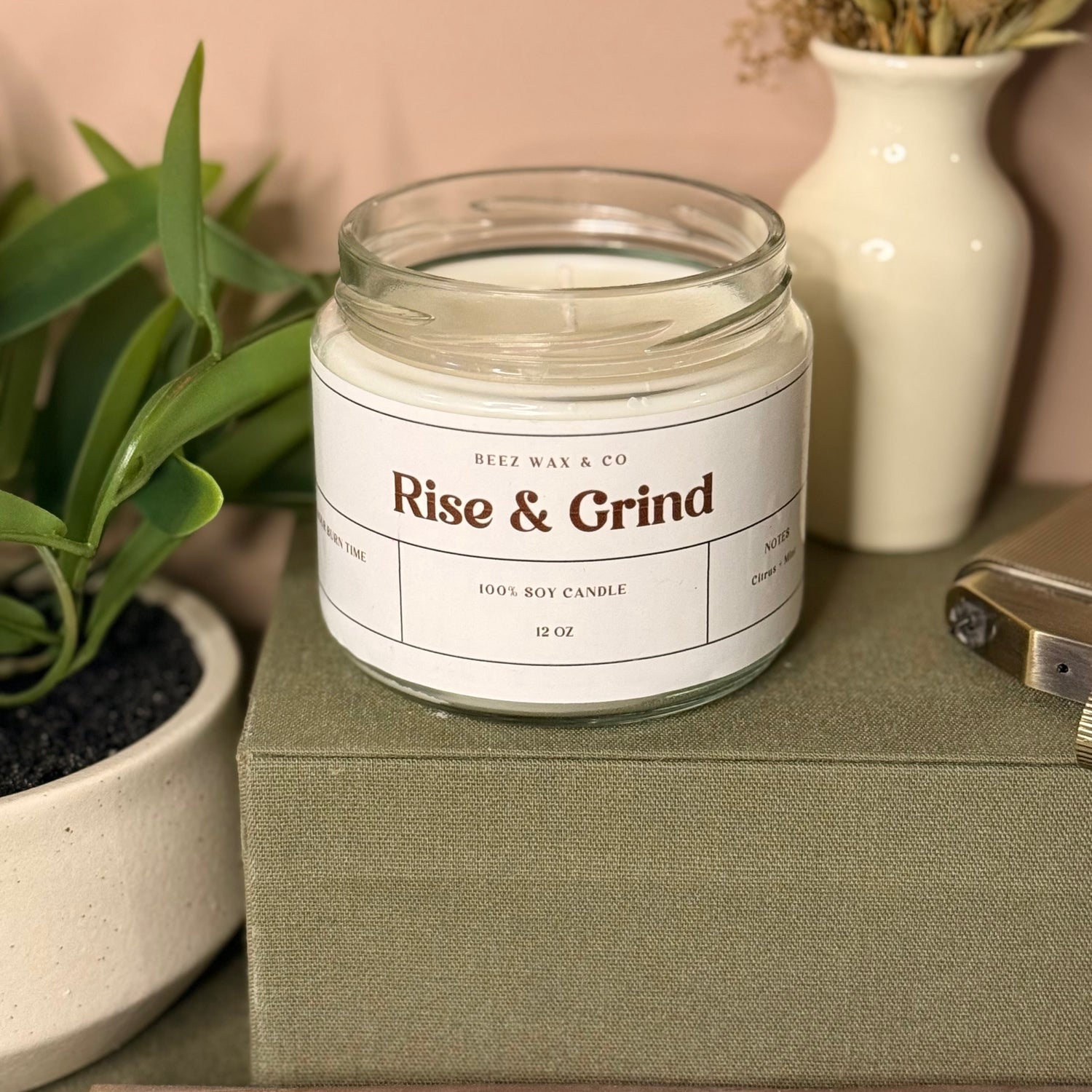 Candle labeled 'Rise & Grind' on a surface with a plant and vase in the background