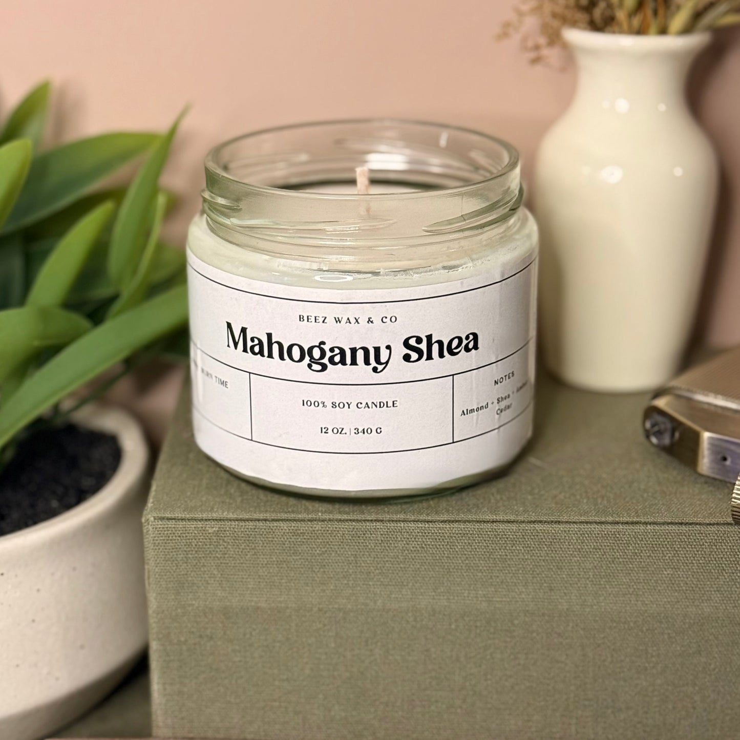 Candle labeled 'Mahogany Shea' on a green box with a plant and vase in the background