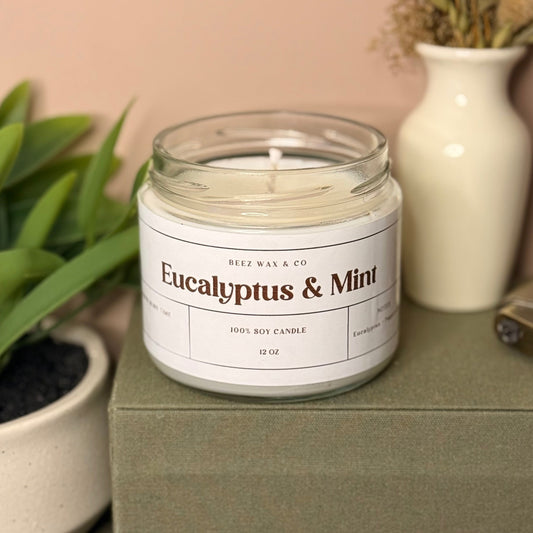 Candle labeled 'Eucalyptus & Mint' on a surface with plants and a vase in the background.