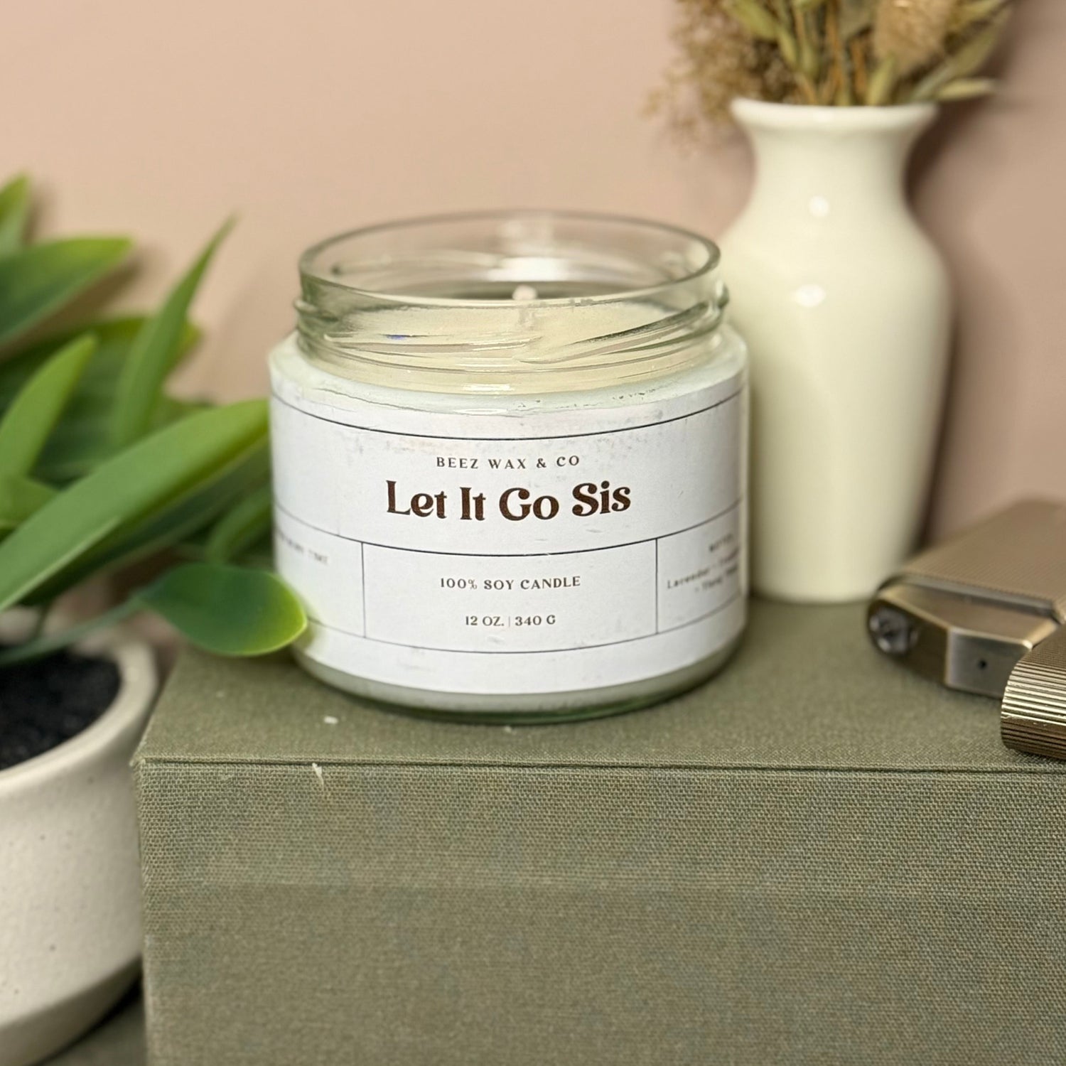 Jar of candle labeled 'Let It Go Sis' on a surface with a plant and vase in the background.