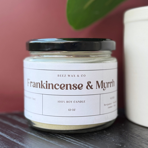 Jar of 'Frankincense & Myrrh' candle from Beez Wax & Co on a wooden surface with a plant in the background.
